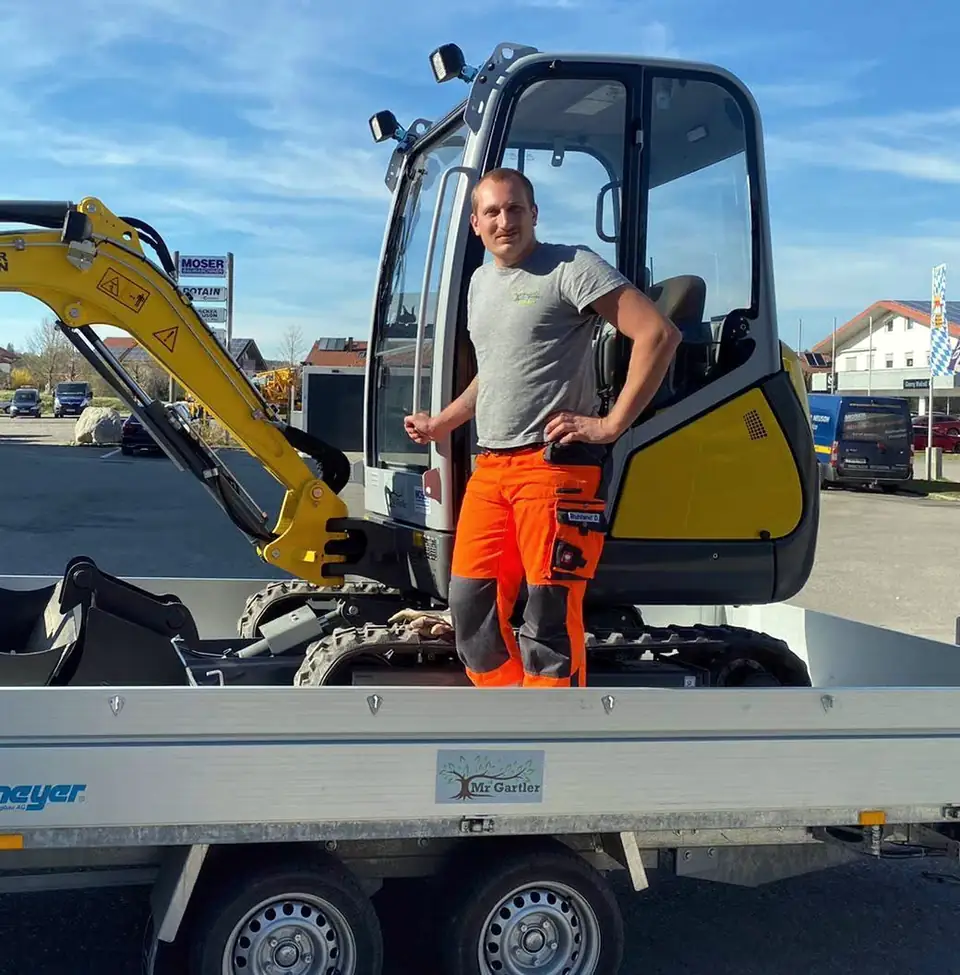 Dominik Ruhland steht auf dem Mr.-Gartler-Anhänger neben einem Wacker-Neuson-Minibagger
